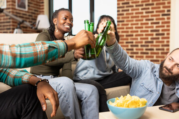 Happy multiethnic friends clink beer bottles and sitting on couch in modern brick wall apartment. Cheerful group enjoys drinks, snacks and fun moments during casual home gathering.