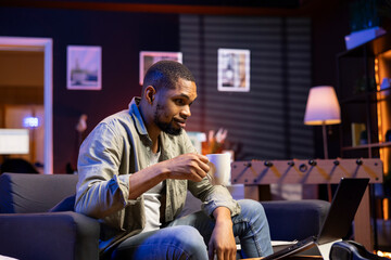 African american smiling man sipping on coffee working remotely, finishing his tasks at the coffee table in the living room. Casual relaxed person browsing the internet and multitasking at home.