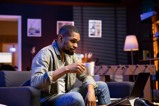 African american smiling man sipping on coffee working remotely, finishing his tasks at the coffee table in the living room. Casual relaxed person browsing the internet and multitasking at home.