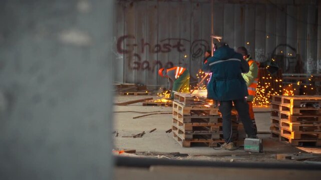 Builders in protective gear use metal cutters to shape materials. Sparks fly as they work diligently in an empty construction area surrounded by wooden pallets and unfinished structures
