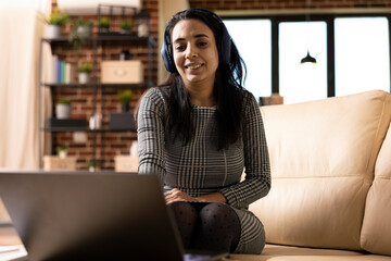 Smiling woman wearing wireless headphones and watching movie on laptop during break from remote work. Indian female freelancer seated on couch, listening to music while searching for online gigs.