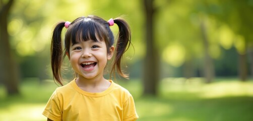 Happy little girl with Down syndrome smiles in park. Portrait of joyful child with pigtails and yellow shirt outdoors at summer. Nature background with green trees.