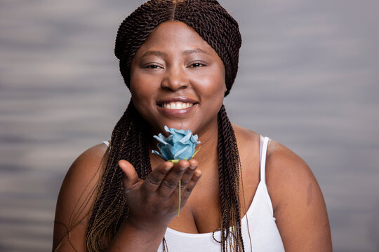 Positive black model posing with a blue rose and radiant smile in studio, advertising natural beauty and self acceptance. Woman with glossy skin and soft complexion does advertisement.