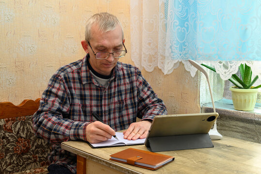 adult man in glasses writing with a pen in a notebook while engaged in online learning at home