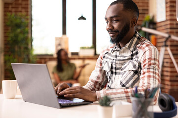 African american male blogger working from home, completing online journal for his website. Black man researching marketing strategies on laptop and gathering data for business project presentation.