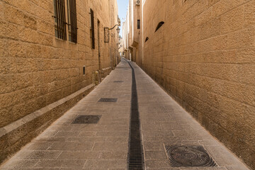 long, narrow alleyway in the Jerusalem Old City is captured, featuring towering walls constructed of rough, golden-hued stone blocks.  winding perspective of the historic passage.
