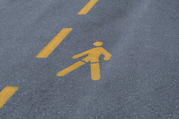 yellow pedestrian symbol is painted onto the dark gray asphalt surface. road pavement also features two parallel, dashed yellow lines, indicating a designated walking lane or crossing zone.