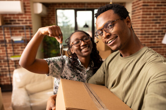 Smiling black man and woman hugging and posing with house keys and cardboard box, marking excitement of first home ownership. Happy couple taking selfie in living room of new apartment.
