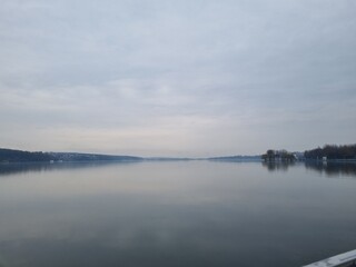 A wide, serene view of a large lake or river with calm water reflecting the dull, flat gray and white clouds of an overcast sky in the early spring