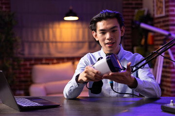 Focused young adult holds virtual reality headset while speaking into microphone, recording promotional video content. Creative media influencer sharing tech insights on vr glasses for online viewers.