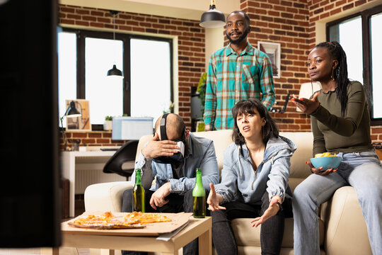 Diverse friends gather at home, reacting to dramatic video game moment. Multiethnic men and women show disappointment while sitting on couch, holding VR headset and controllers during weekend hangout.
