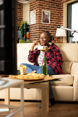 Casual afternoon indoors as young woman eats noodles from takeout box while lounging on sofa. Black lady munching on noodles and watching sports or movie on tv in cozy apartment living room.