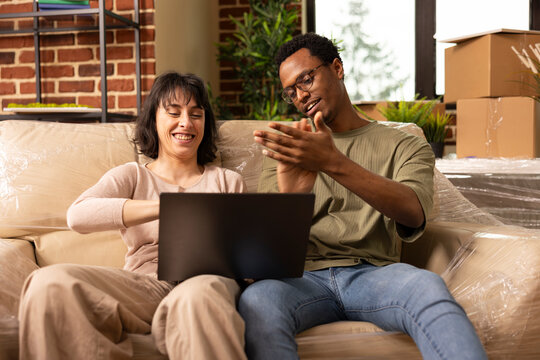 Cheerful white woman looks at device screen as African American husband claps, seated on couch in new apartment. Boyfriend cheering for girlfriend after finding favorite item on laptop for home.