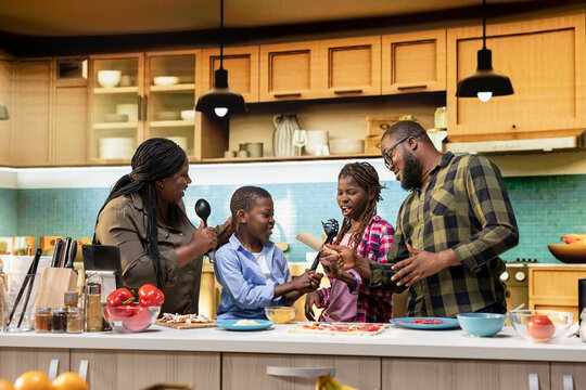 African american young family singing and dancing around in the kitchen, acting silly and laughing together for fun weekend activity. Goofy kids and parents using utensils for karaoke.