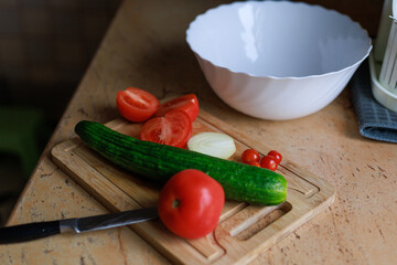 Cucumber, tomatoes and onion on a cutting board near a bowl. Concept of healthy cooking, ingredient prep and simple food styling.