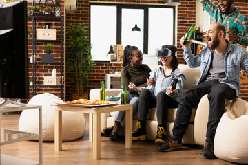 Diverse young adults enjoys online gaming with VR headsets and wireless controllers in cozy living room. Group of friends celebrate friendship with food, drinks and fun during relaxed hangout.