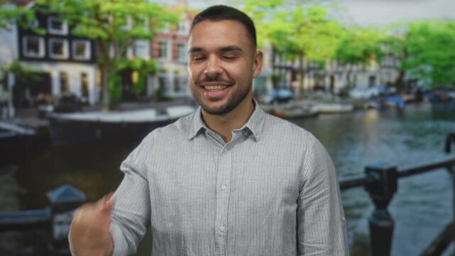 Man wearing light striped shirt shows callme hand gesture with fingers near ear and winks while smiling on street; friendly connection.