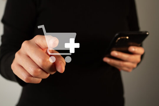 A woman points to a shopping cart on her phone, with a plus sign. The shopping cart is a symbol of online shopping.