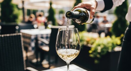 Waiters hand pouring white wine into a glass at an outdoor sunny terrace, blurred summer restaurant background, refreshing vibe, focus on the bottle neck and flow