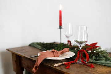 Stylish christmas table setting for family dinner. Elegant plate with napkin, cutlery, glassware, ribbons, green fir branches, red berries and candle on wooden table. Christmas Holiday time