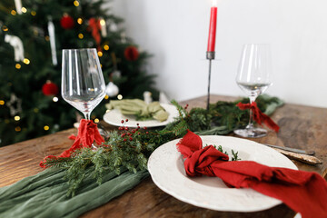 Stylish christmas table setting with modern plate, red and green napkins, cutlery, glass, fir branches, red berries, candle on wooden table. Merry Christmas and Happy Holidays!