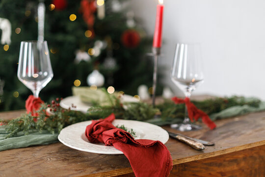 Stylish christmas table setting with modern plate, red and green napkins, cutlery, glass, fir branches, red berries, candle on wooden table. Merry Christmas and Happy Holidays!