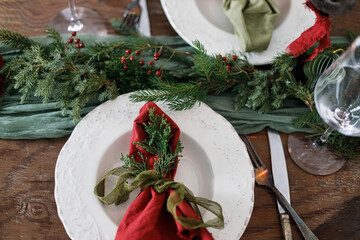 Merry Christmas! Stylish table setting for christmas family dinner. Modern plate and cutlery with napkin, glassware, green and red ribbons, fir branches and candle on rustic table flat lay