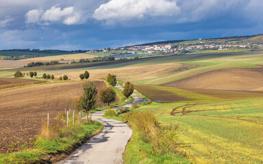 Obraz premium The country of South Moravia in the Czech Republic, Europe. Picturesque landscape featuring rolling hills, a winding road, and a distant town under a cloudy sky. Serene country charm revealed.