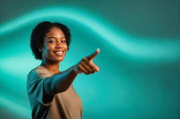 Fototapeta premium A smiling woman with short dark hair pointing forward, set against a teal-colored wavy light backdrop in a studio setting.