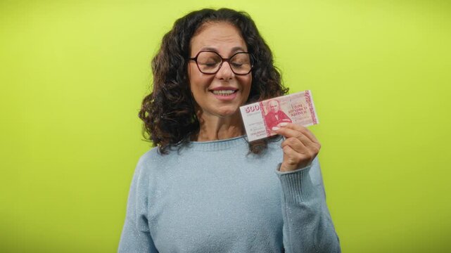Middle-aged woman shows icelandic krona banknote with playful expression against a vibrant yellow background, highlighting the concept of finance and currency with a touch of humor.