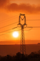 A towering electricity pylon silhouette stands majestically against a blazing orange sky at sunset. Power lines stretch across a scenic, rural landscape.