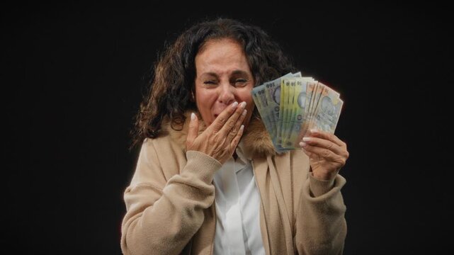 Middle-aged woman joyously holding romanian banknotes against isolated black background, expressing happiness and surprise in her cozy sweater and lei.