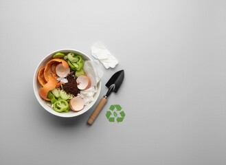 Composting kitchen waste: Vegetable peels, eggshells, coffee grounds, and paper scraps in a white bowl with a garden trowel and green recycling symbol on a light gray background with copy space