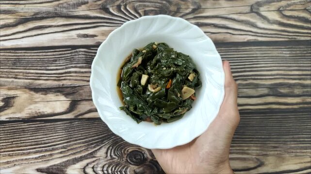 Hands holding a white plate of stir-fried genjer vegetables. Genjer vegetables, Limnocharis flava, Sawah-lettuce, or yellow velvetleaf, are aquatic plants that can be cooked into dishes.