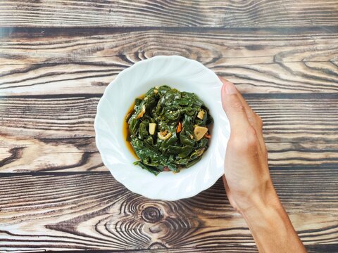 Hands holding a white plate of stir-fried genjer vegetables. Genjer vegetables, Limnocharis flava, Sawah-lettuce, or yellow velvetleaf, are aquatic plants that can be cooked into dishes.