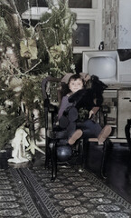 A vintage portrait of a sweet little girl sitting in an antique chair and holding a teddy bear near a Christmas tree. Retro photo from 1972.