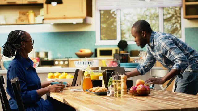 African american man in a hurry running late for his office job, skipping breakfast and leaving his housewife to eat alone at the table. Boyfriend waving goodbye and rushing to leave. Camera B.