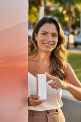A smiling woman holding a white rectangular product box and pointing at it, standing outdoors in natural light with a blurred green background.