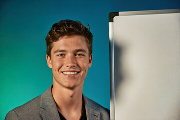 A young man with short brown hair and a friendly smile stands in front of a whiteboard against a teal gradient background, wearing a gray blazer over a black shirt.