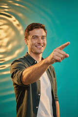 A smiling young man in a dark shirt pointing to the side against a teal and gold abstract background.