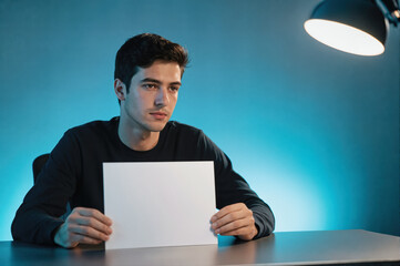 A young man sitting at a desk under a lamp, holding a blank white paper in front of him against a blue-lit background.