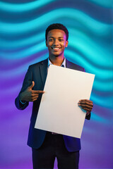 A smiling young man in a dark suit holding a blank white board and pointing at it, standing against a vibrant blue and purple wavy background.