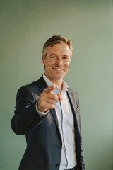 A smiling man in a dark suit and light blue shirt pointing directly at the camera against a solid green backdrop.