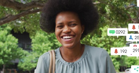 Smiling woman standing under trees in park, wearing sage-green blouse and tan strap, app icons