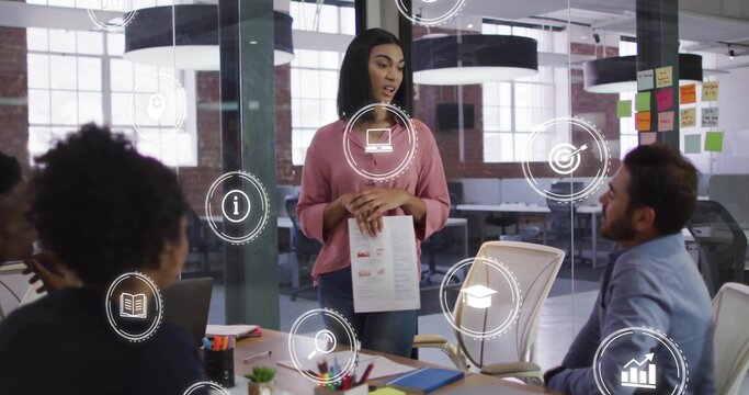 Presenting adult woman in pink pullover and jeans holding report in open-plan office, with laptops