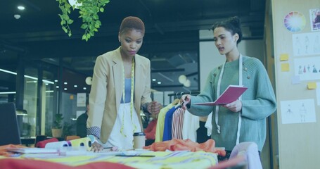Two women designers examining fabrics in studio, in blazer and sweater holding measuring tape