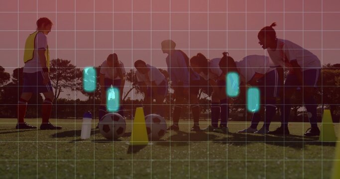 Practicing team and coach stretching on grassy pitch at sunset, with balls, cones, yellow bib, grid
