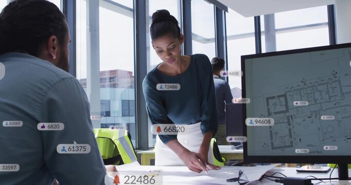 Leaning architect reviewing plans at office table, wearing dark blouse white skirt, with monitor