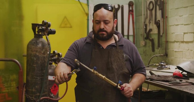 Inspecting bearded welder in dark apron checking oxy-fuel torch valves in shop, with gas cylinders - Powered by Adobe