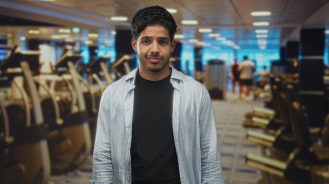 Man points thumbs to chest in gym amid treadmills and cardio machines wearing open shirt and black t shirt; casual confidence.